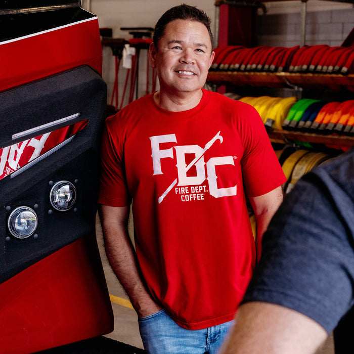 A man wearing a red Fire Dept. Coffee T-shirt featuring a large white FDC logo with a pike pole graphic running through the middle and small ‘Fire Dept. Coffee’ text below, standing beside a red fire truck inside a fire station with coiled hoses in the background
