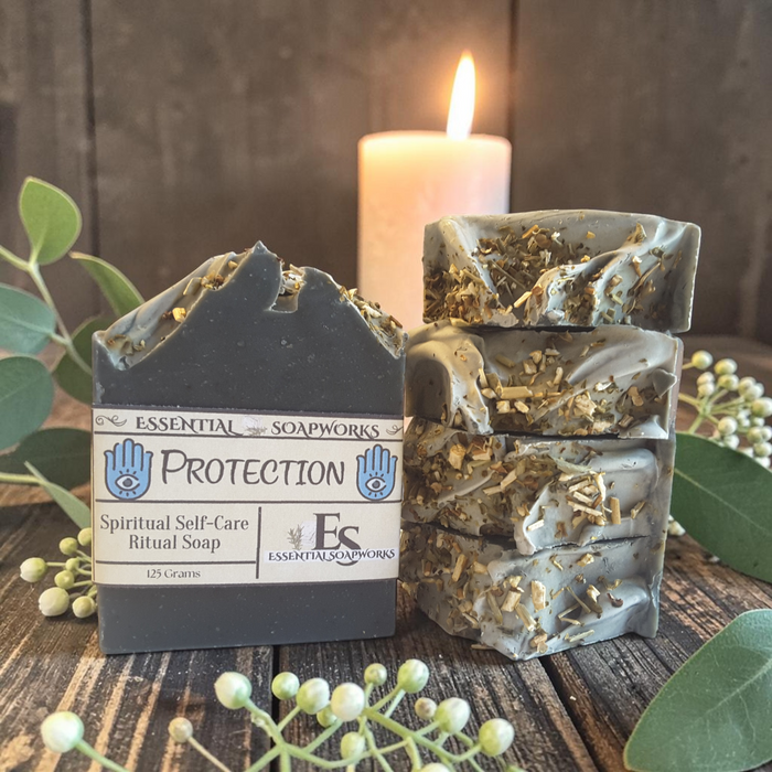 A bar of soap labeled 'Protection' by Essential Soapworks stands next to a stack of four matching bars. The soap is dark charcoal gray with dried botanicals embedded in it, styled on a rustic wooden surface with a lit candle, green leaves, and small white berries in the background.