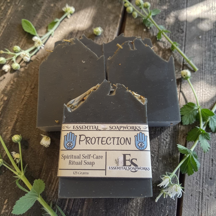 Two dark charcoal gray handmade soap bars with jagged, rustic edges, one wrapped in a paper label that reads 'PROTECTION Spiritual Self-Care Ritual Soap' by 'Essential Soapworks'. The soaps are arranged on a rustic dark wood background decorated with green leaves and small white flowers.
