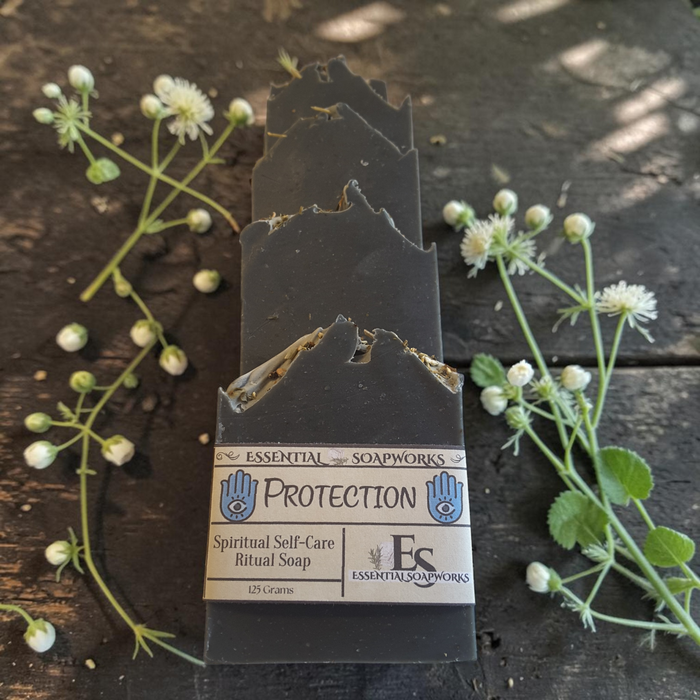 A rectangular bar of dark charcoal ritual soap with a jagged, rustic edge and a paper label reading 'Protection' by Esoteric Soapworks, placed on a dark wooden surface with small white flowers around it.