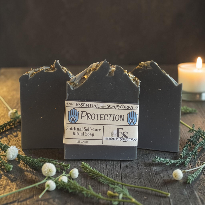 Three handmade rectangular bars of soap with jagged, rustic tops are arranged on a dark wooden surface. The center bar is upright and shows a paper label reading 'Essential Soapworks Protection Spiritual Self-Care Ritual Soap,' while the two side bars are leaning behind it.