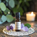A small amber glass bottle labeled 'Psychic Awareness Ritual Oil' sits on a white lace doily with purple crystals and green leaves, with a blurred lit candle in the background.