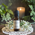 A small amber glass bottle of Purification Ritual Oil with a black cap and a cream label sits on a white lace doily, surrounded by white flowers and soft green foliage, with a blurred lit candle in the background.