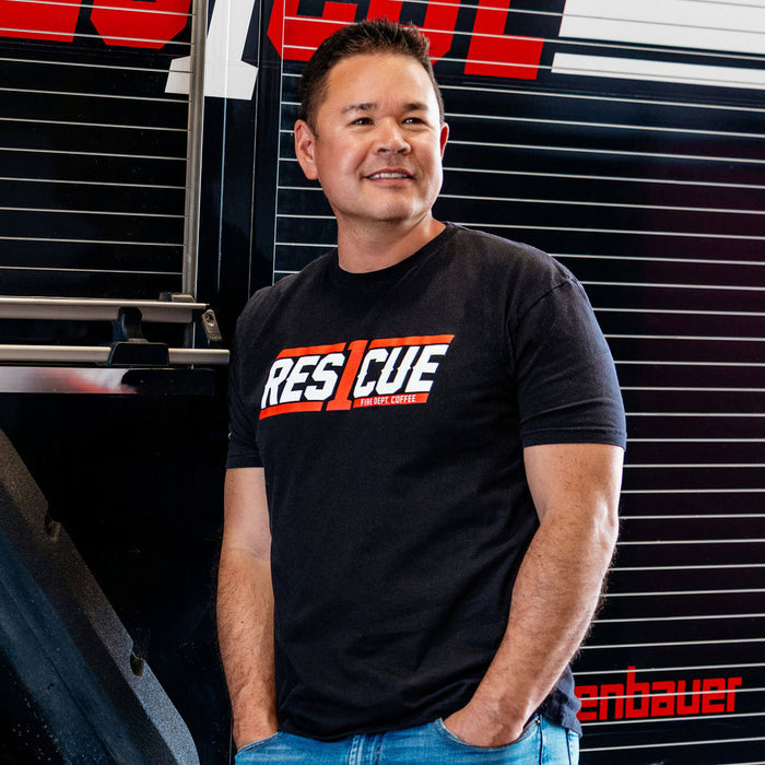 A man standing next to a fire truck wearing the Rescue1 Shirt, featuring the Rescue1 logo.