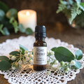 A small amber glass bottle of Road Opener Ritual Oil stands on a white lace doily, surrounded by eucalyptus leaves and white berries, with a blurred lit candle in the background.