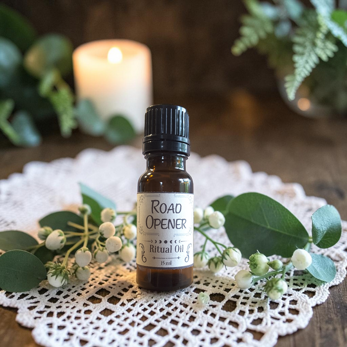 A small amber glass bottle of Road Opener Ritual Oil stands on a white lace doily, surrounded by eucalyptus leaves and white berries, with a blurred lit candle in the background.