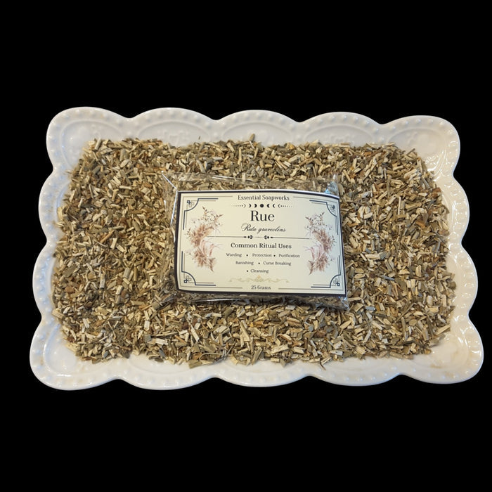 A square white ceramic dish with a scalloped edge holds loose dried rue with a clear plastic bag and printed label centered on top. The label reads 'Rue'.