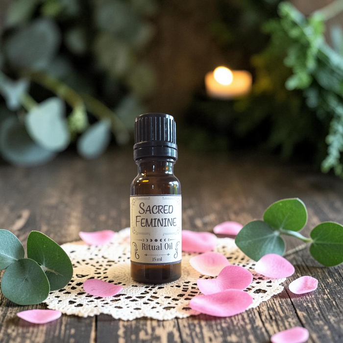 A small amber glass bottle of Sacred Feminine ritual oil with a black cap, styled on a wooden surface with a lace doily, pink petals, and green eucalyptus leaves. The background is a soft-focus, warm, natural setting with a glowing candle.
