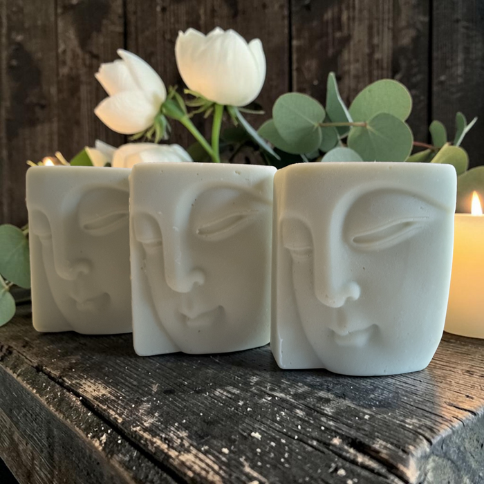 three bars of buddha face shaped soap on a wooden surface with white flowers and candles in the background