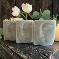 three bars of buddha face shaped soap on a wooden surface with white flowers and candles in the background