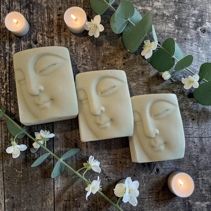 Three rectangular soap bars with embossed serene face designs, arranged on a rustic wooden surface with lit tea lights, white flowers, and green eucalyptus leaves.