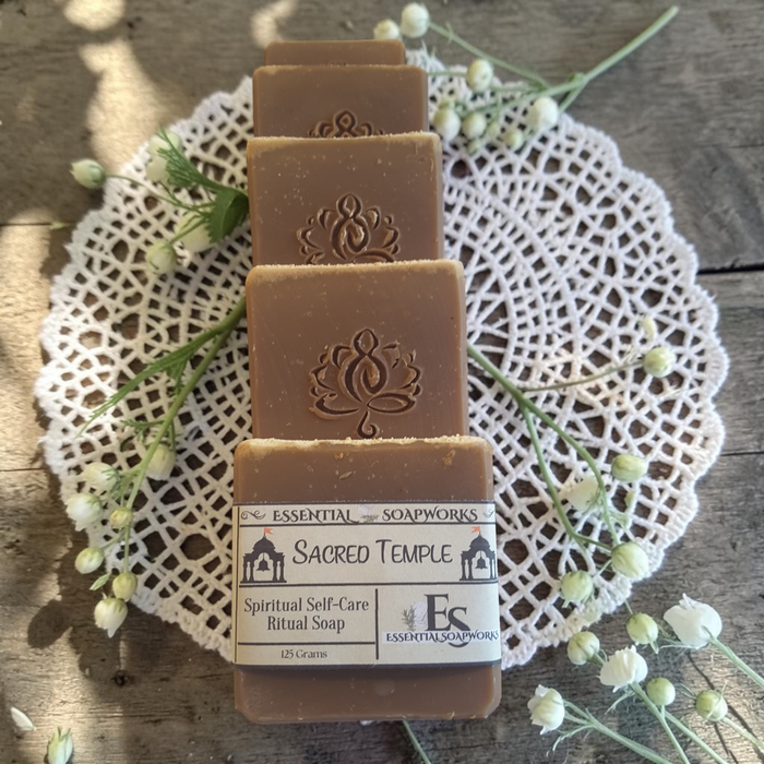 Four rectangular bars of brown ritual soap stacked vertically on a white lace doily over a rustic wooden surface. Each bar is embossed with a lotus symbol, and the bottom bar has a paper label reading 'ESSENTIAL SOAPWORKS SACRED TEMPLE Spiritual Self-Care Ritual Soap 120 Grams'.