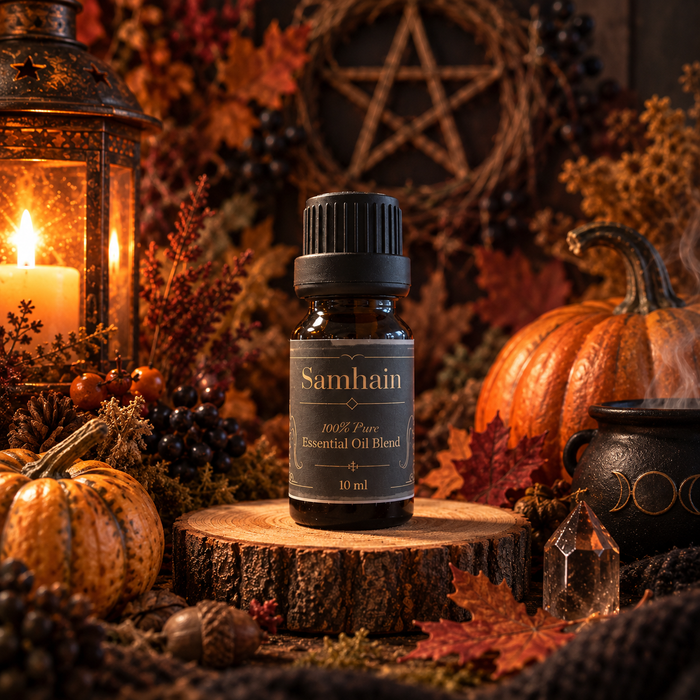 A 10 ml bottle of Samhain 100% Pure Essential Oil Blend displayed on a rustic wood slice. The setting is a cozy, atmospheric autumnal arrangement featuring a lit candle in a lantern, pumpkins, a cauldron, and autumn leaves.