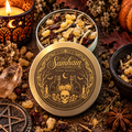 A round metal tin of Samhain Loose Incense is shown open with the lid in front, surrounded by autumn herbs, crystals, dried flowers, a candle, and spices on a rustic surface. The lid features a detailed gold illustration of cats, pumpkins, a skull, and a crescent moon on a black background, with the words 'Samhain Loose Incense' printed above.