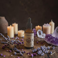 A bottle of Samhain Ritual Oil stands on a wooden surface surrounded by lit candles, lavender sprigs, dried petals, and purple crystal stones against a dark rustic background.