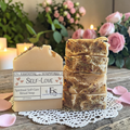 four bars of self love soap on a doily on a wooden surface with roses and candles in the background
