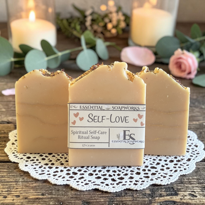 Three bars of Essential Soapworks 'Self-LOVE' spiritual self-care ritual soap displayed on a white lace doily over a rustic wooden surface, with blurred lit candles, eucalyptus leaves, and a pink rose in the background.