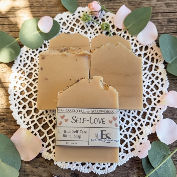 Three rectangular bars of tan ritual soap with visible botanical inclusions, arranged on a white lace doily over a rustic wooden surface with green eucalyptus leaves and pale pink petals. The front bar has a paper label reading 'ESSENTIAL SOAPWORKS SELF-LOVE Spiritual Self-Care Ritual Soap'.