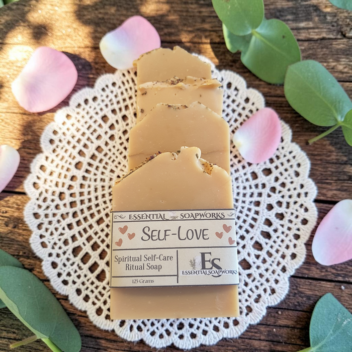 Four bars of Essential Soapworks 'Self-Love' Spiritual Self-Care Ritual Soap are arranged on a white lace doily over a rustic wooden surface, surrounded by green leaves and pink petals.