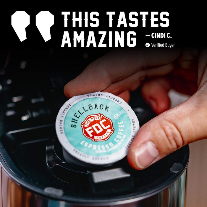 A hand inserts a round coffee capsule into a black coffee maker. The capsule label reads 'SHELLBACK' and 'FOC Specialty Coffee', with a large promotional text overlay at the top that says 'THIS TASTES AMAZING' and 'CINDI C.