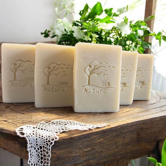 Five bars of '100% Natural' soap on a wooden surface with plants in the background.