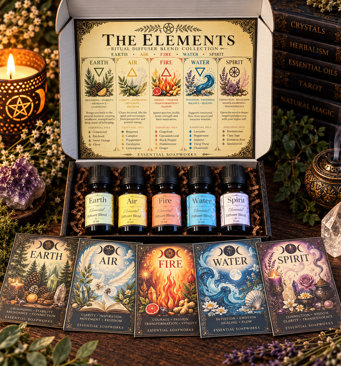 A boxed set of five essential oil diffuser blend bottles labeled Earth, Air, Fire, Water, and Spirit, displayed open with five matching illustrated cards in front. The set is arranged on a dark wooden surface with candles, crystals, and books around it.