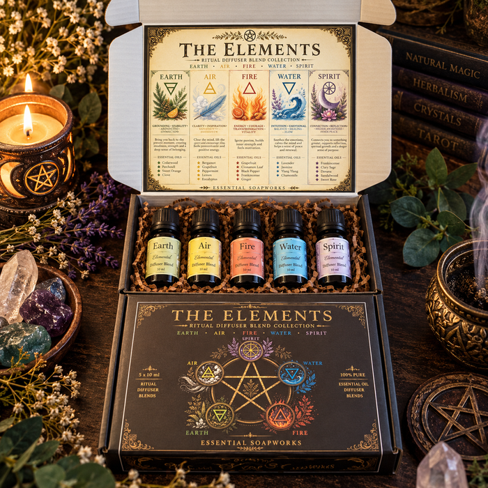 An open gift box containing five small ritual diffuser blend bottles labeled Earth, Air, Fire, Water, and Spirit, with the branded lid and front box visible. The box is styled on a dark wooden surface with candles, crystals, dried flowers, and books around it.