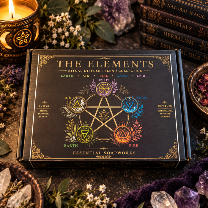 The Elements Ritual Diffuser Blend Collection box by Essential Soapworks, featuring a black box with gold lettering and a pentagram design, surrounded by crystals, a lit candle, and books on a dark rustic surface.