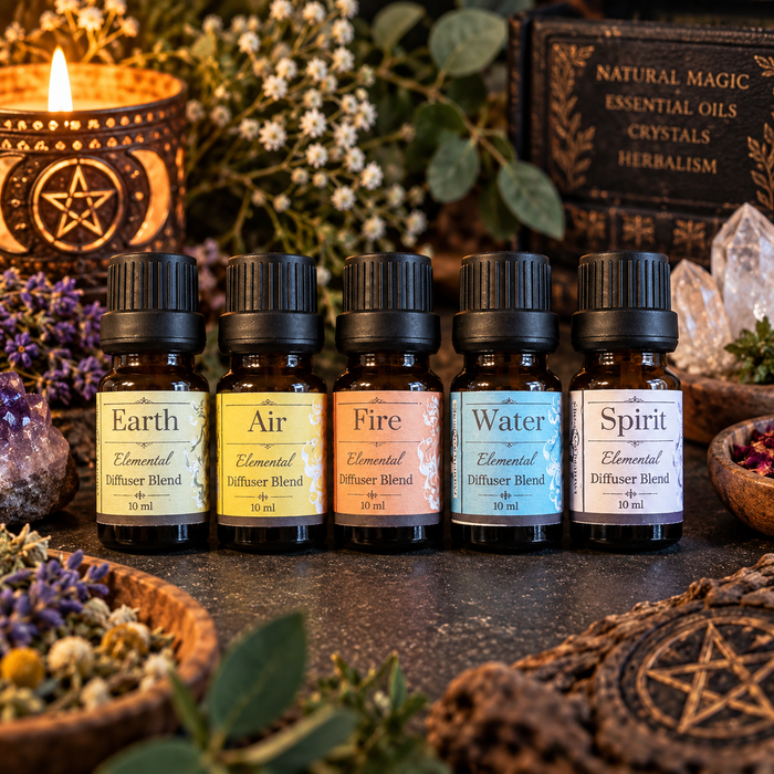 Five small amber glass essential oil bottles labeled Earth, Air, Fire, Water, and Spirit are lined up on a dark tabletop with candles, herbs, crystals, and a book in the background.