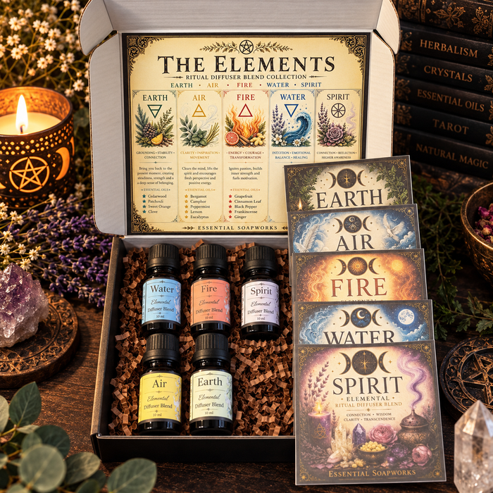 An open gift box of 'The Elements' essential oils and diffusers set, showing five small bottles and five illustrated cards, styled on a wooden surface with candles, crystals, and books.