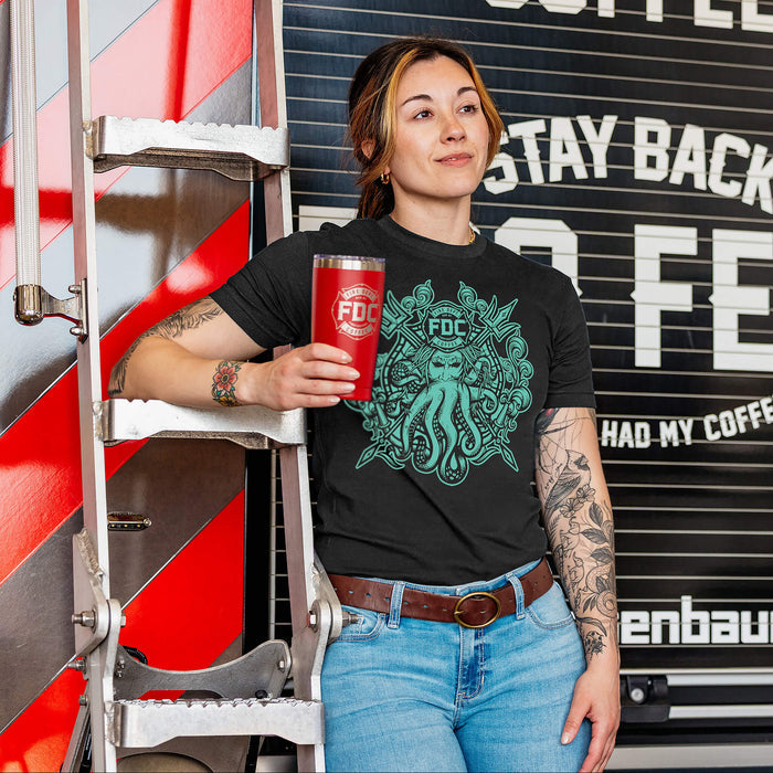 A woman wearing a black Fire Dept. Coffee T-shirt featuring a large teal Shellback-inspired graphic with an FDC logo and octopus design on the front, leaning beside a fire truck ladder while holding a red Fire Dept. Coffee tumbler,  and a fire truck in the background
