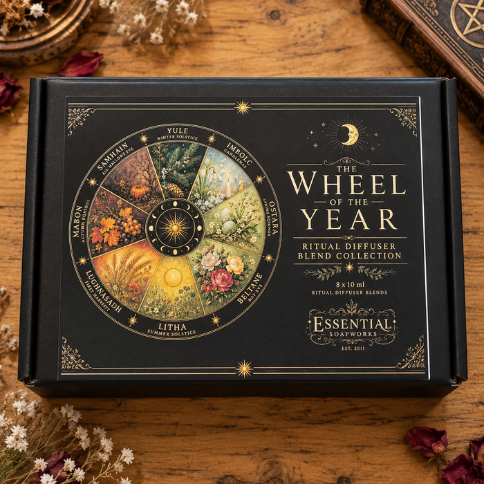 A black gift box labeled 'The Wheel of the Year Ritual Diffuser Blend Collection' by Essential Essences, placed on a wooden surface with dried flowers and a brass wheel nearby.