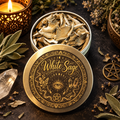 A round metal tin of dried white sage with its lid placed in front, decorated with an ornate gold-toned eye and pentagram design labeled 'White Sage.' The tin is styled on a dark surface with dried leaves, a lit candle, crystals, and a small pentacle charm.