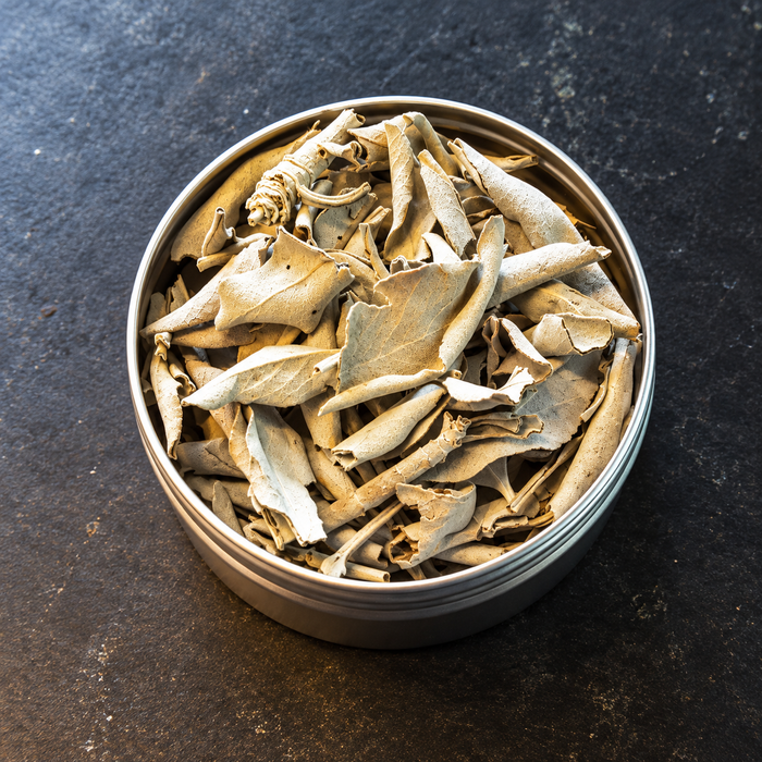 Dried white sage inside a round metal tin, photographed from a top-down view on a dark textured surface.