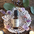 A small amber glass bottle labeled 'Witch's Brew Ritual Oil' sits centered on a white lace doily, surrounded by purple crystals, dried lavender, green leaves, and candles on a rustic wooden surface.