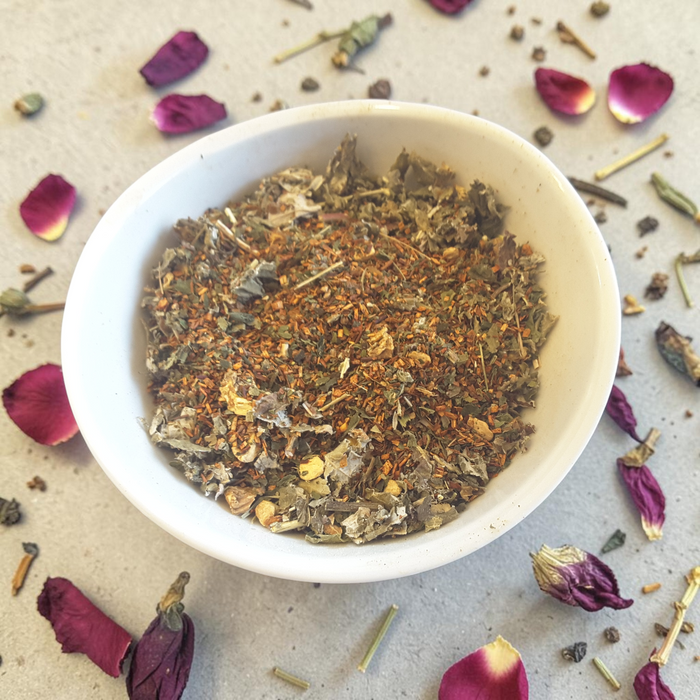 loose leaf sacred feminine tea blend i a white bowl on a light surface with scattered petals and herbs.