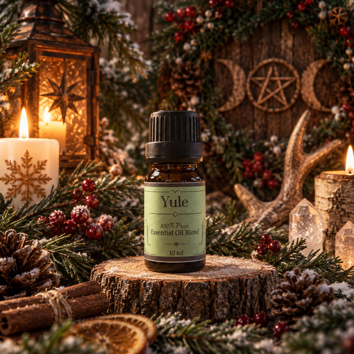 A 10 ml amber glass bottle of Yule Essential Oil Blend with a black cap and a light green label, displayed on a rustic wood slice surrounded by pine branches, candles, pinecones, and winter decorations.
